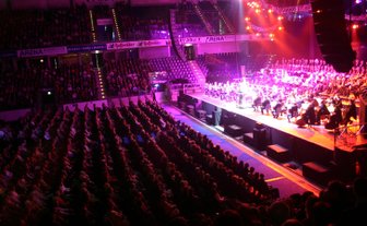 Ein großes Publikum sitzt in einem Konzertsaal, während ein Orchester auf der Bühne spielt, umgeben von bunten Lichtern.