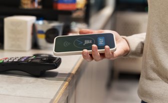 Eine Person hält ein Smartphone mit einem NFC-Symbol, bereit zur kontaktlosen Zahlung an einem Kassenterminal.