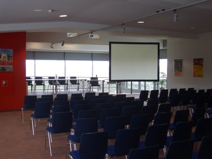 Ein moderner Konferenzraum mit Reihen von blauen Stühlen und einer großen Leinwand im Hintergrund, bereit für eine Veranstaltung.