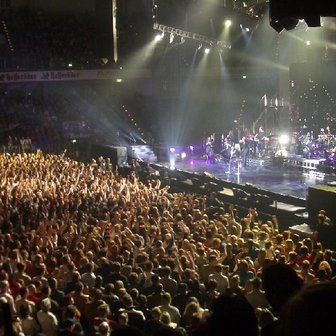 Eine große Menschenmenge jubelt begeistert bei einem Konzert, während die Band auf der Bühne spielt und Lichter strahlen.