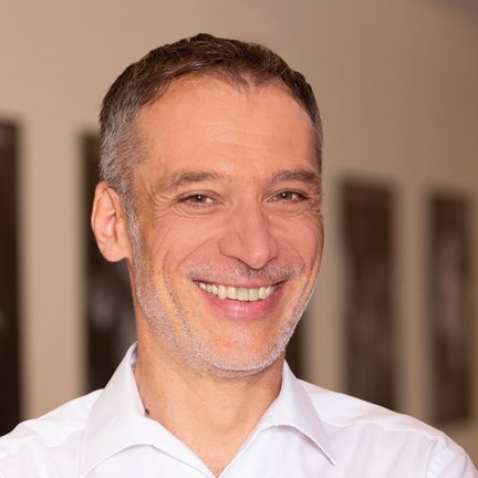 Ein lächelnder Mann mit grauen Haaren trägt ein weißes Hemd und steht vor einer neutralen Hintergrundwand.