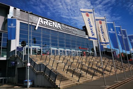 Die Außenansicht der Arena zeigt große Glasfenster und markante Treppen mit Handläufen, umgeben von Fahnen.