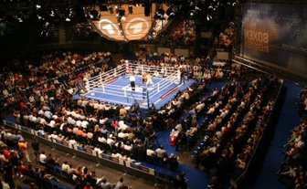 Eine große Menschenmenge beobachtet einen Boxkampf in einer beleuchteten Arena mit einem blauen Boxring in der Mitte.