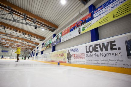 Eine Eishalle mit mehreren Werbebannern an der Wand, während Schlittschuhläufer auf dem Eis ihre Runden drehen.