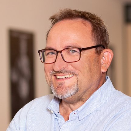 Ein lächelnder Mann mit Brille trägt ein hellblaues Hemd und steht in einem modernen Innenraum.