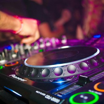 Das Mischpult eines DJs wird von einer Hand bedient, während im Hintergrund eine pulsierende Partystimmung mit bunten Lichtern herrscht.