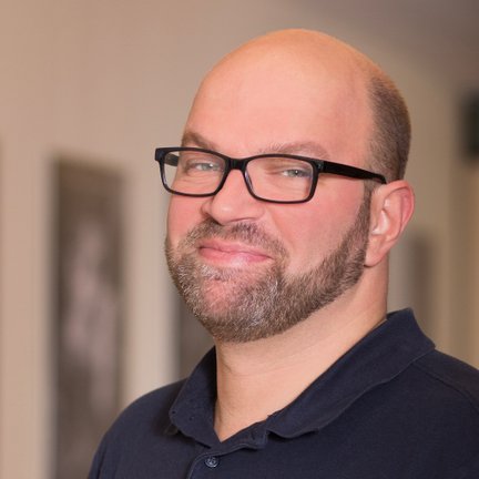 Ein lächelnder Mann mit Brille und Bart steht in einem hellen Raum, umgeben von unscharfen Bildern an der Wand.