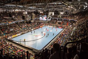 Ein spannendes Handballspiel in einer vollbesetzten Halle, mit Spielern in roten und schwarzen Trikots, die um den Ball kämpfen.