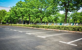 Ein weitläufiger Parkplatz mit leeren Stellplätzen, umgeben von üppigem Grün und Bäumen unter einem klaren blauen Himmel.