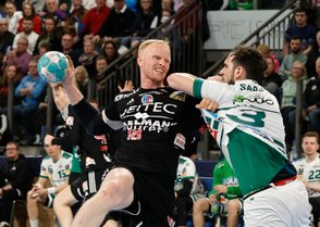 Ein Handballspieler in schwarzer Kleidung wirft den Ball, während er von einem Gegner in Weiß angegriffen wird.