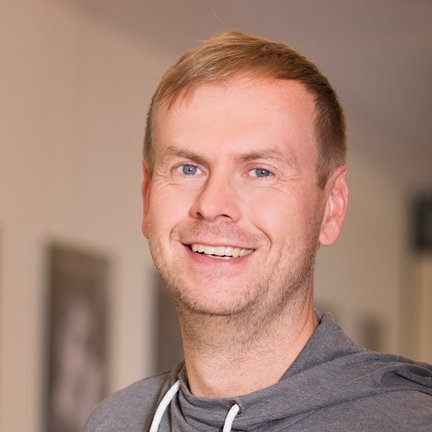 Ein lächelnder Mann mit blonden Haaren trägt einen grauen Pullover und steht in einem hellen Raum mit Bildern im Hintergrund.