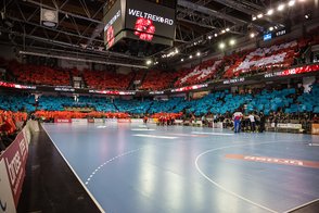 Ein Handballspiel in einer Arena, die mit bunten Tüchern in Rot und Blau dekoriert ist, während die Spieler sich versammeln.