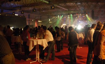 Die Veranstaltung zeigt eine große Gruppe von Personen, die in einem hell erleuchteten Raum mit rotem Teppich feiern und tanzen.