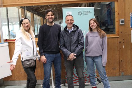 Eine Gruppe von vier Personen lächelt fröhlich vor einem Holz-Hintergrund, während sie sich in einer Sporteinrichtung befinden.