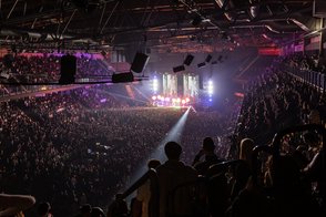 Eine große Menschenmenge genießt ein Konzert in einer Arena, umgeben von bunten Lichtern und einer lebhaften Atmosphäre.