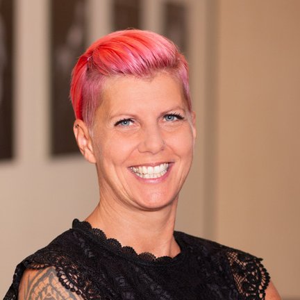 Mit einem selbstbewussten Lächeln präsentiert sich die Frau mit pinken Haaren in einem stilvollen, schwarzen Kleidungsstück.