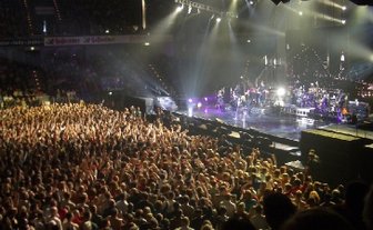 Eine große Menschenmenge jubelt begeistert bei einem Konzert, während die Band auf der Bühne spielt und Lichter strahlen.