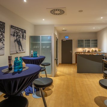 Der Innenraum zeigt stilvolle Stehtische mit blauen Tischdecken, umgeben von Hockeykunstwerken und einer einladenden Bar.
