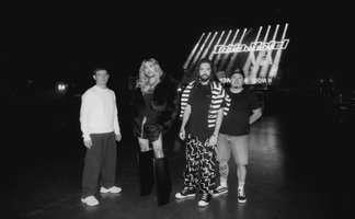 Die Bandmitglieder posieren in modischen Outfits, während im Hintergrund ein großes Bühnenbanner sichtbar ist.