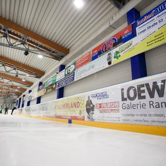 Eine Eishalle mit mehreren Werbebannern an der Wand, während Schlittschuhläufer auf dem Eis ihre Runden drehen.