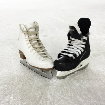 Auf dem glatten Eis liegen ein weißer und ein schwarzer Eishockeyschuh, die Schnürsenkel sind ordentlich gebunden.