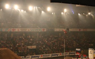 Ein Motocross-Fahrer springt über eine Rampe in einer vollen Arena, umgeben von jubelnden Zuschauern und grellem Licht.