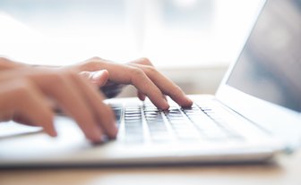 Nahaufnahme von Händen, die schnell auf einer Laptop-Tastatur tippen, umgeben von einem hellen, einladenden Raum.