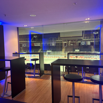 Ein stilvoller Lounge-Bereich mit Möbeln und in abgedunkelt-blauen Farbtönen, der eine klare Sicht auf das Eisstadion und die Zuschauerplätze ermöglicht.
